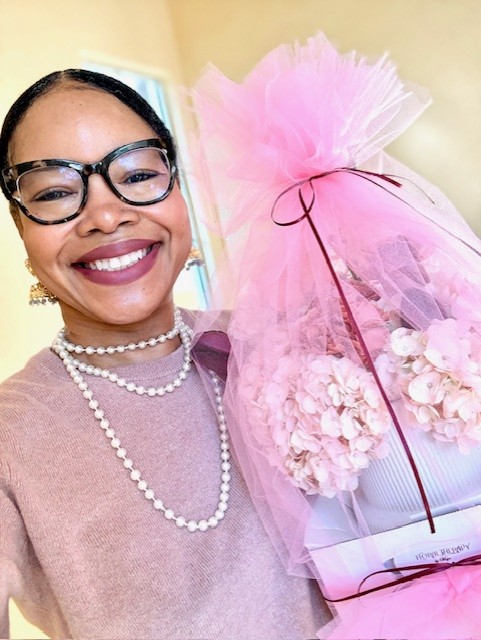 Smiling woman with glasses and pearl necklace holding a pink gift bag with flowers.