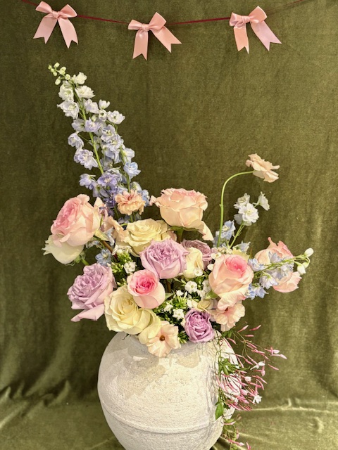 Flower arrangement with pink, purple, and white flowers in a white vase against a green background with pink bows above.