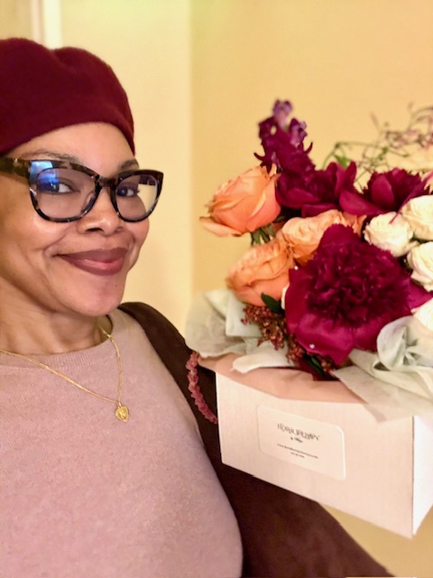 Woman wearing glasses and a red hat holding a bouquet of mixed flowers in a white box.