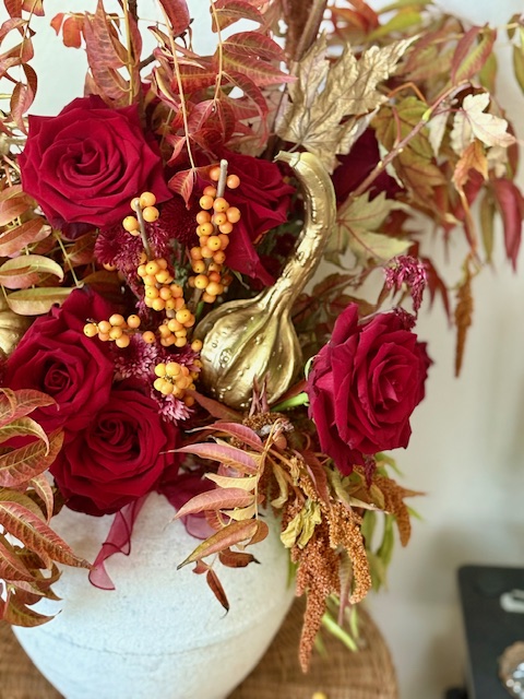 Arrangement of red roses, yellow berries, and autumn leaves in a white vase with a gold decorative element.