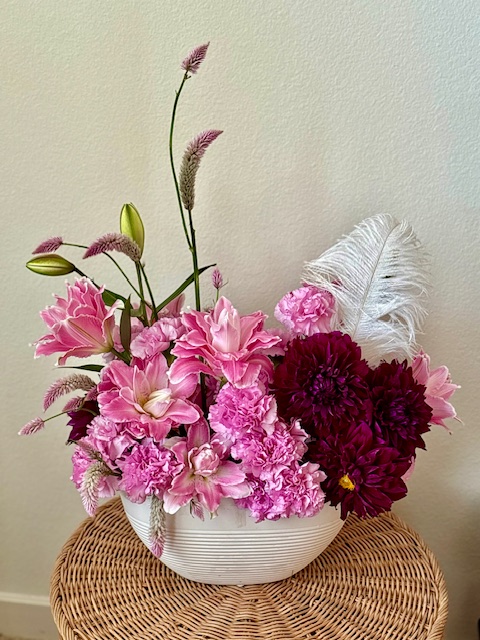 Pink and dark red flowers in a white bowl on a woven surface against a plain wall.