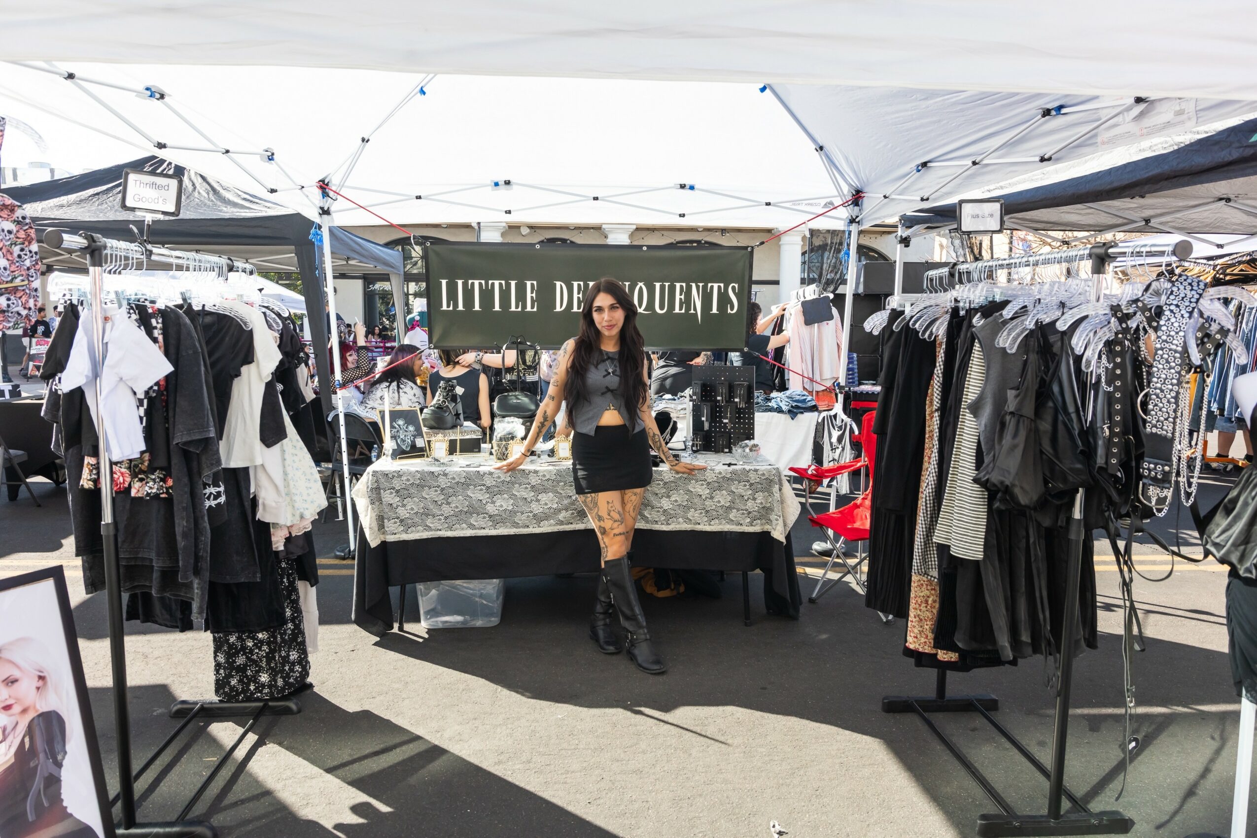 Woman standing in front of a clothing stall at an outdoor market, with racks of clothes on both sides and a sign behind.
