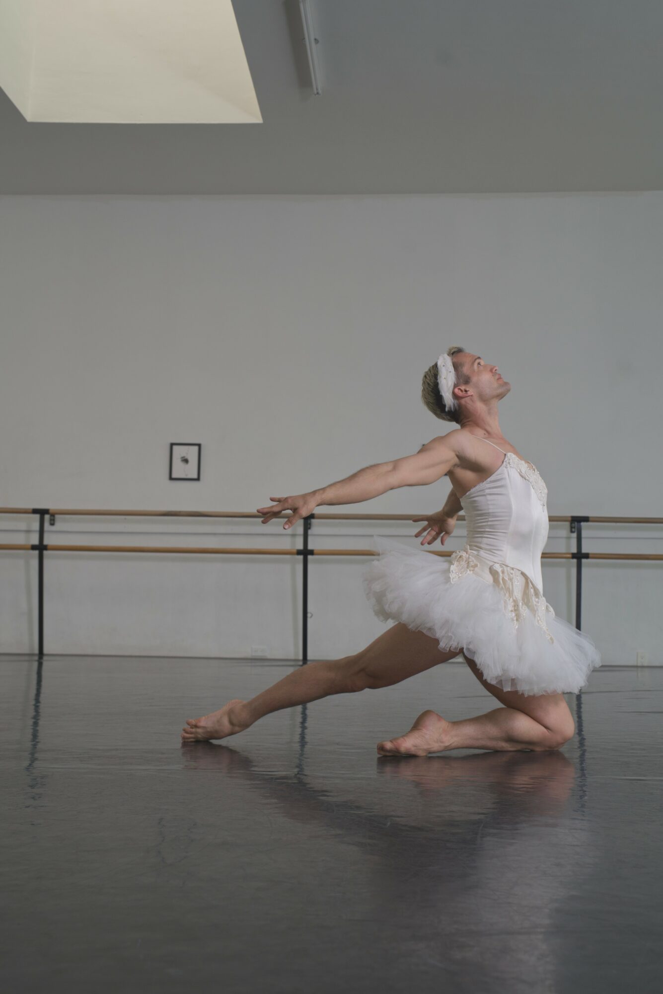 Ballet dancer in white tutu and headband performing a pose in a dance studio with mirrors and bars.