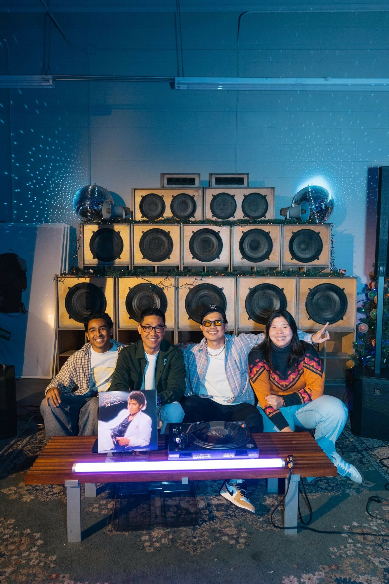 Four people sitting at a table with a large speaker setup behind them, colorful lighting, and a person on a poster in front.