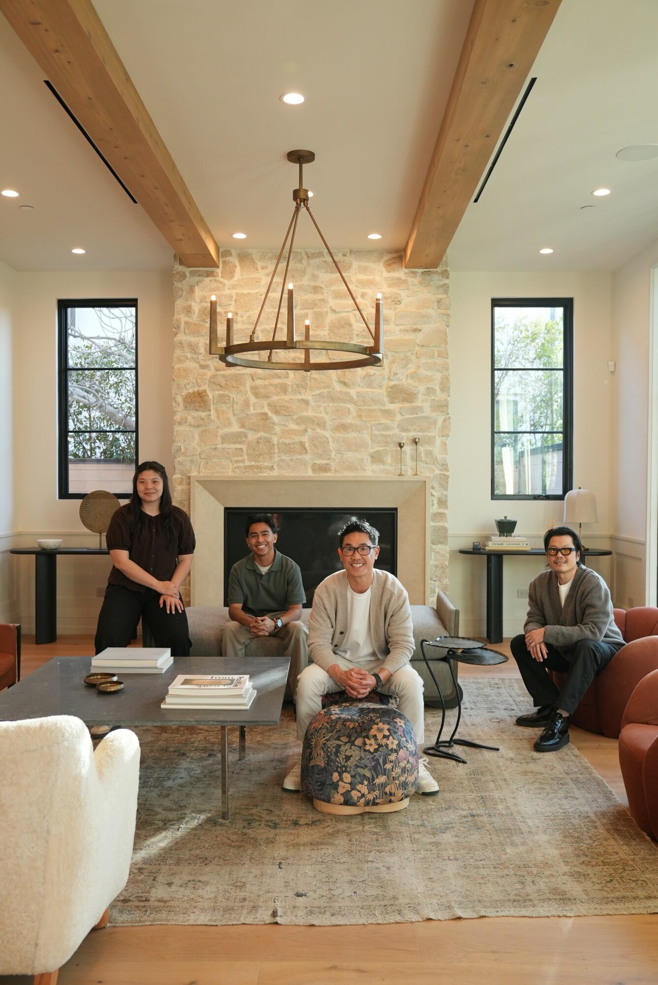 Four people sit and stand in a living room with large windows, a stone fireplace, and a chandelier, with a coffee table.