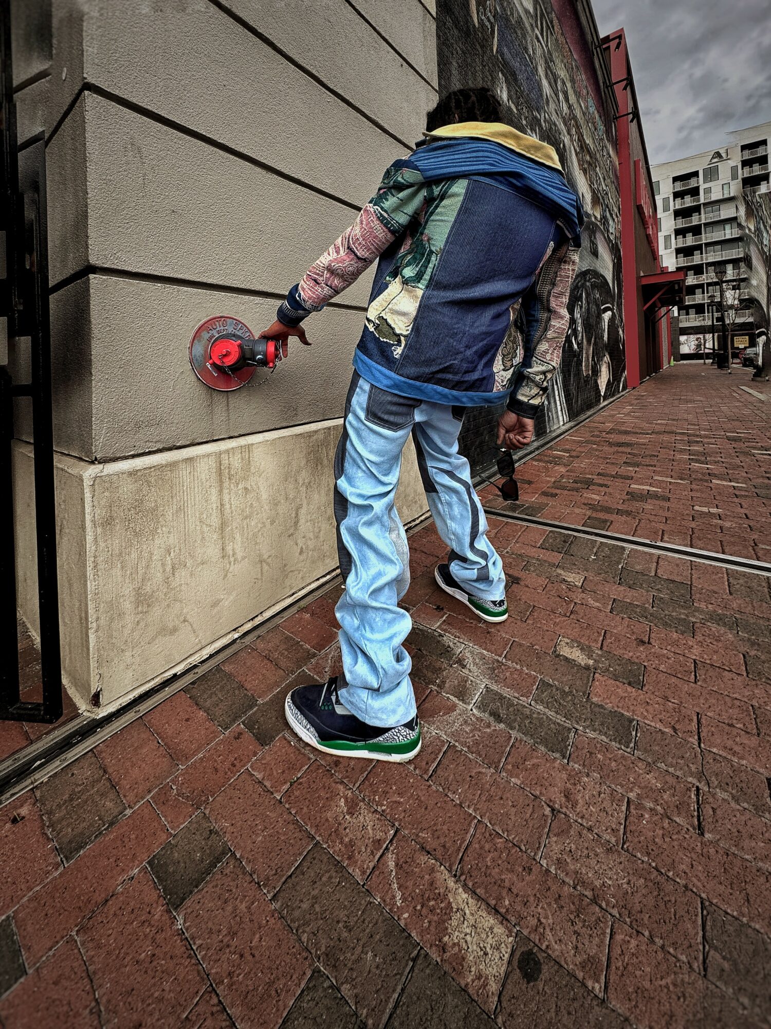 Person using a power tool on a building wall outdoors, wearing a multicolored jacket and jeans.