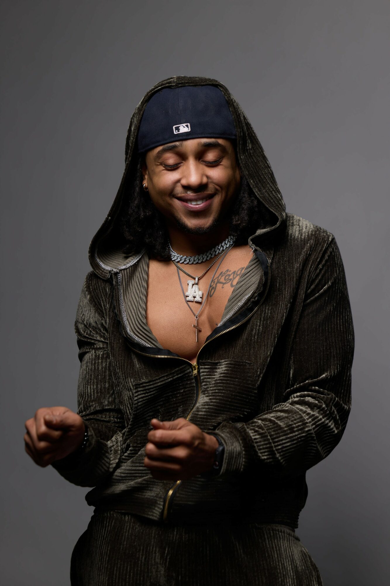 Person with long hair, wearing a cap backwards, smiling, dressed in a dark jacket with a necklace, against a gray background.