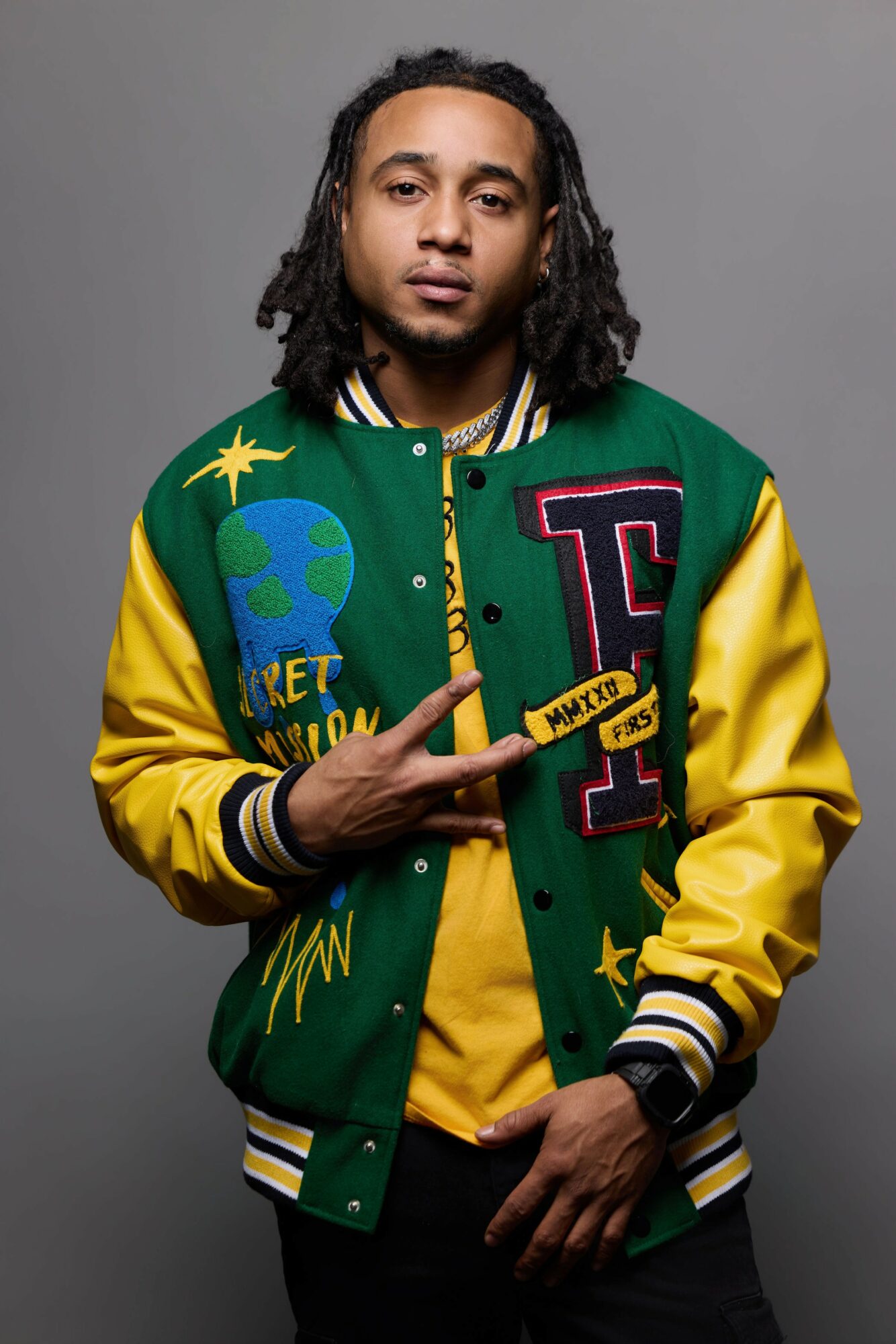 Young man with dreadlocks wearing a colorful varsity jacket, standing against a gray background.