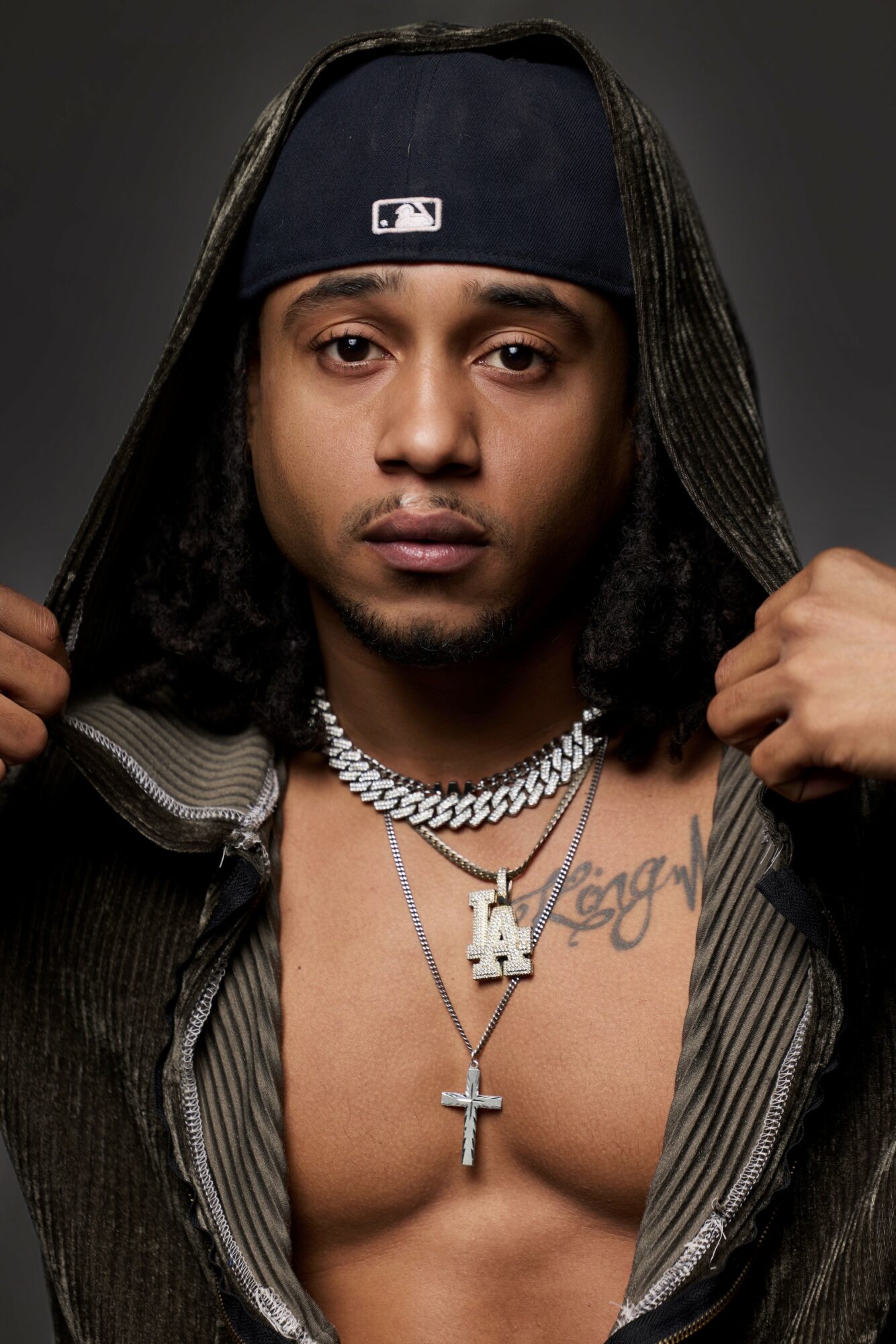 Young man with long hair, wearing a baseball cap, open shirt, and layered necklaces, exposing chest tattoos.