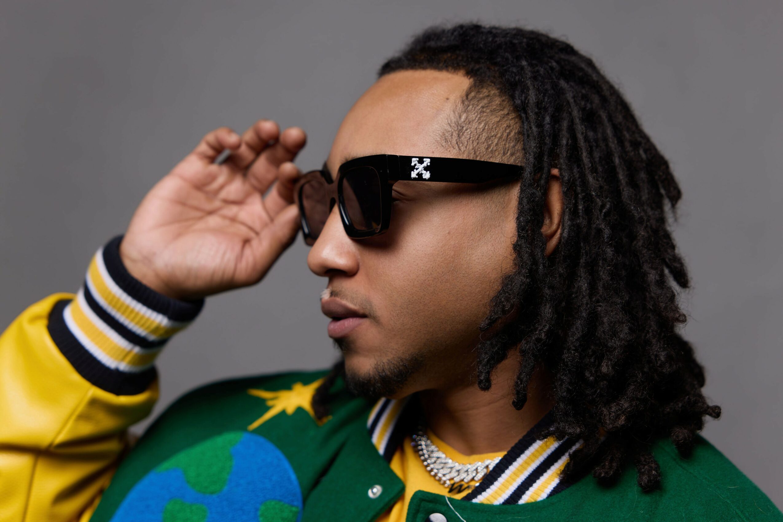Person with dreadlocks wearing sunglasses and a green jacket with yellow and white striped collar, adjusting sunglasses.