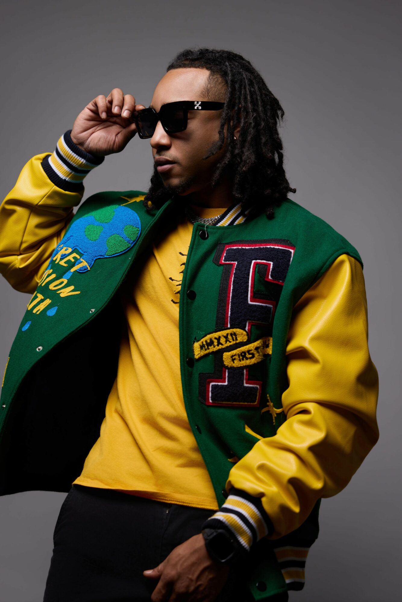 Person wearing sunglasses, green and yellow varsity jacket, yellow shirt, and black pants, posing against a gray background.
