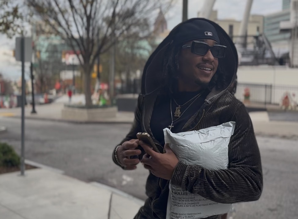 Person wearing sunglasses and a hoodie, holding a bag and a phone, smiling outdoors in an urban setting.