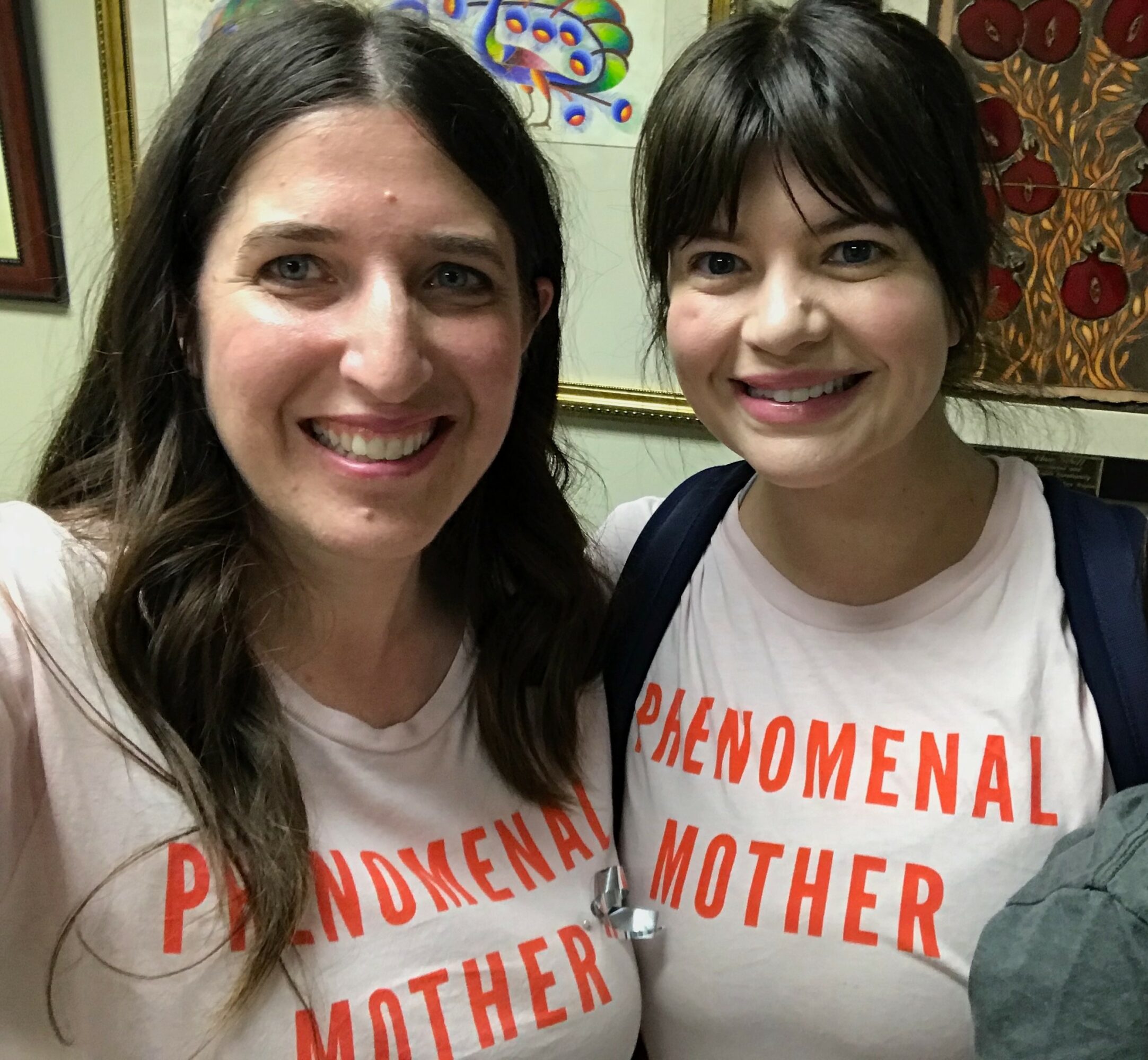 Two women smiling, wearing matching white T-shirts with red text, standing indoors with framed artwork behind them.