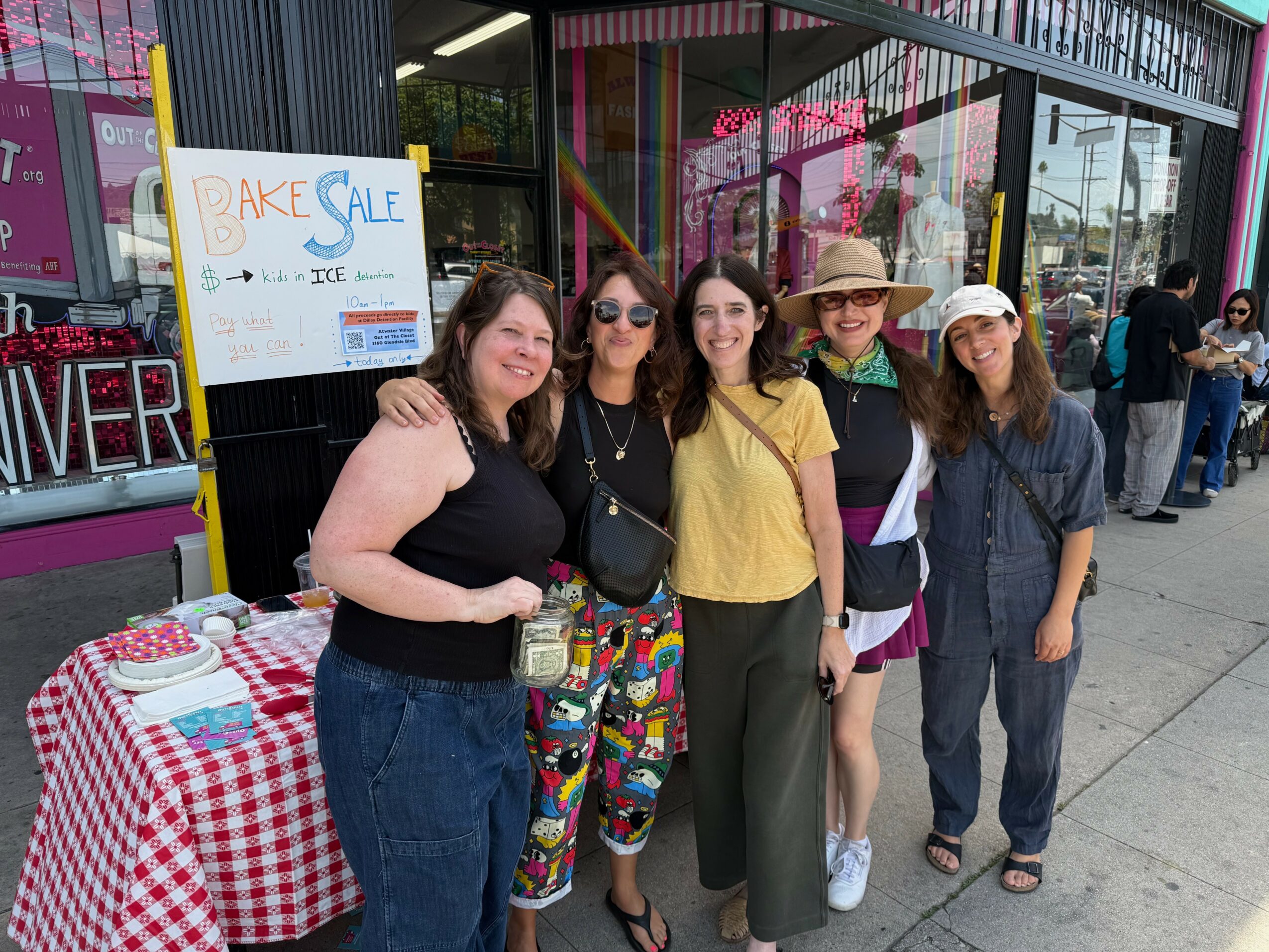 Group of five women standing together outside a store, smiling, with a table and sign nearby.