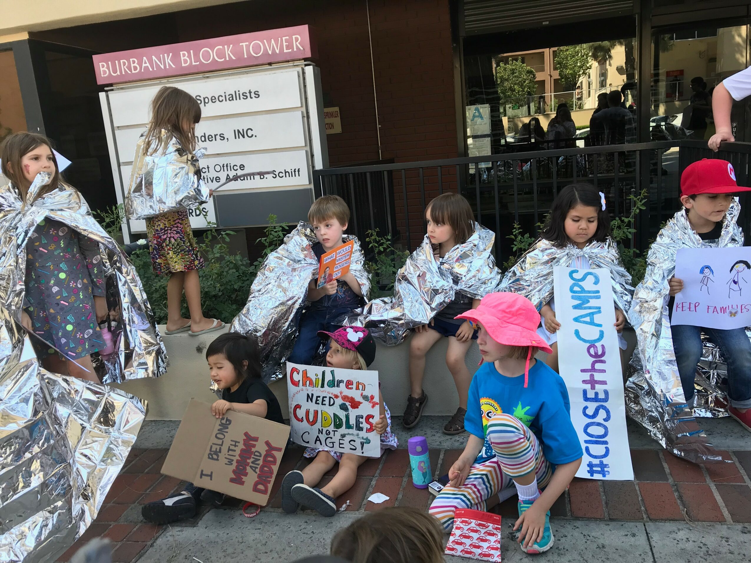 Children sit on the sidewalk holding signs and wearing backpacks, with some standing and others sitting, near a building.