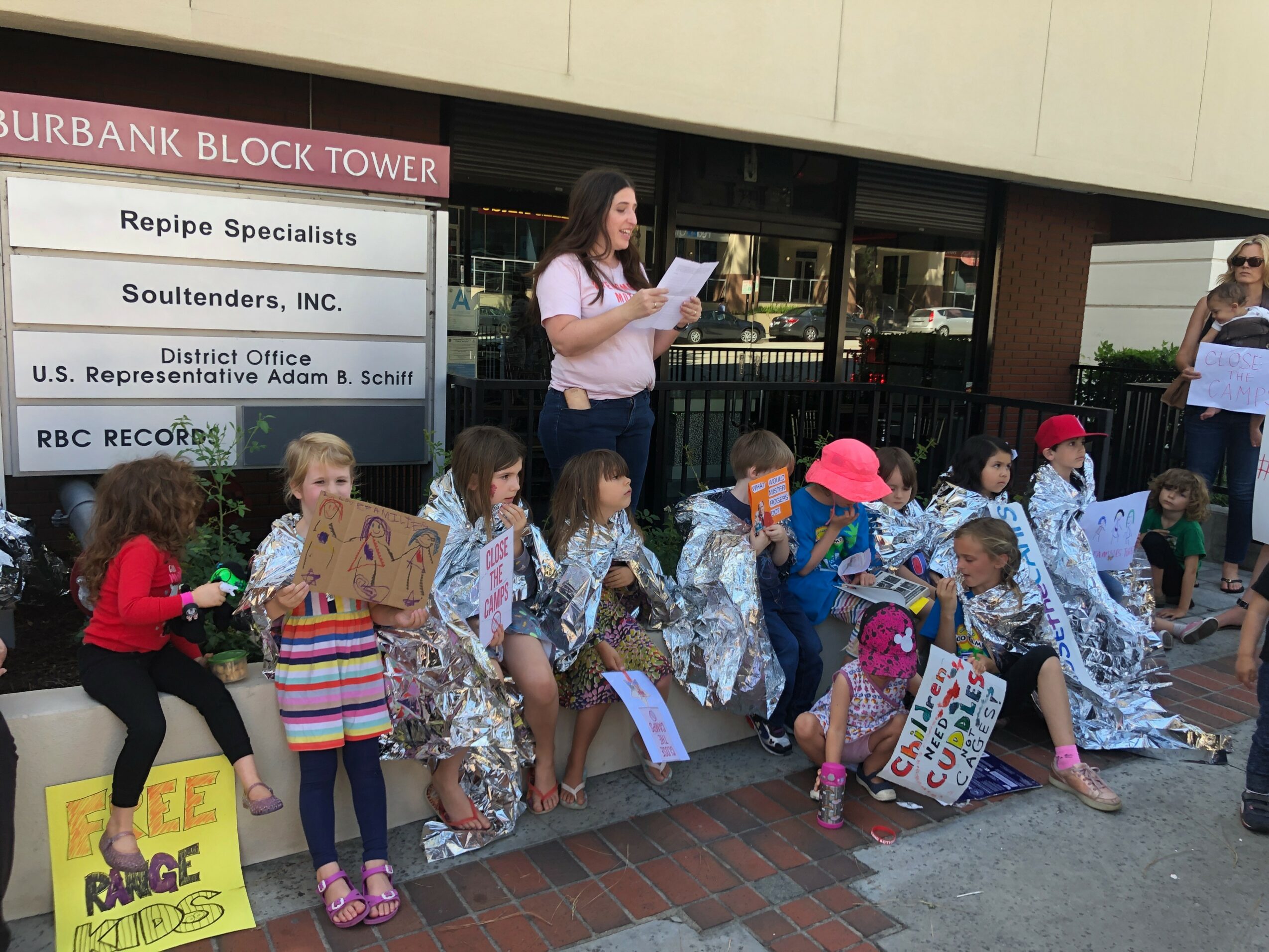 Group of children sitting on a sidewalk, some holding signs and wearing colorful clothing, with adults nearby.