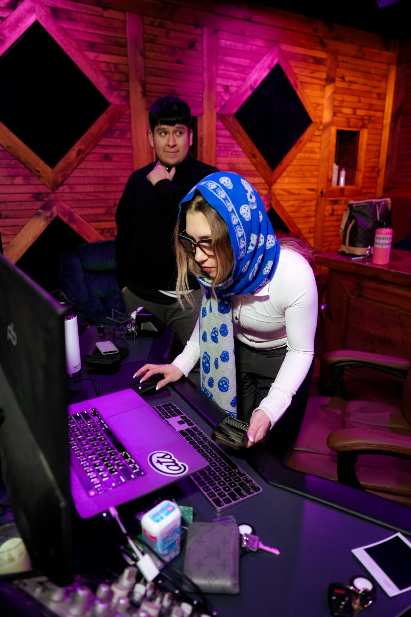 Two women in a room with wooden walls and purple lighting, one working on a computer, the other standing behind.