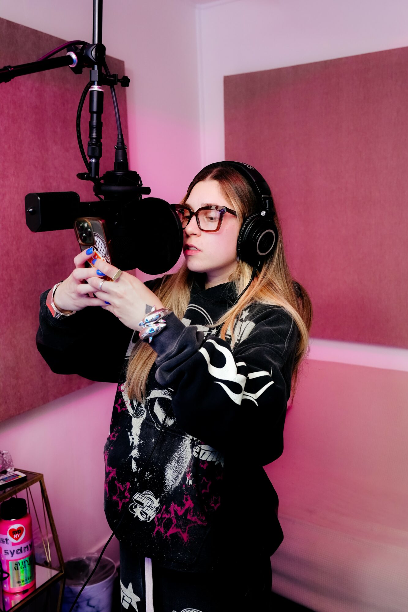 Young woman with glasses and headphones in recording studio, holding a phone, microphone in front, pink wall background.