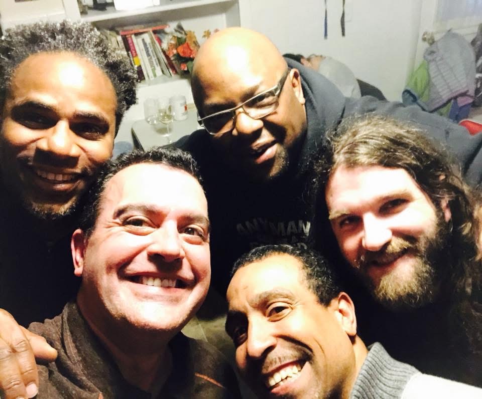 Group of five smiling men in a room with books and clothing in the background.