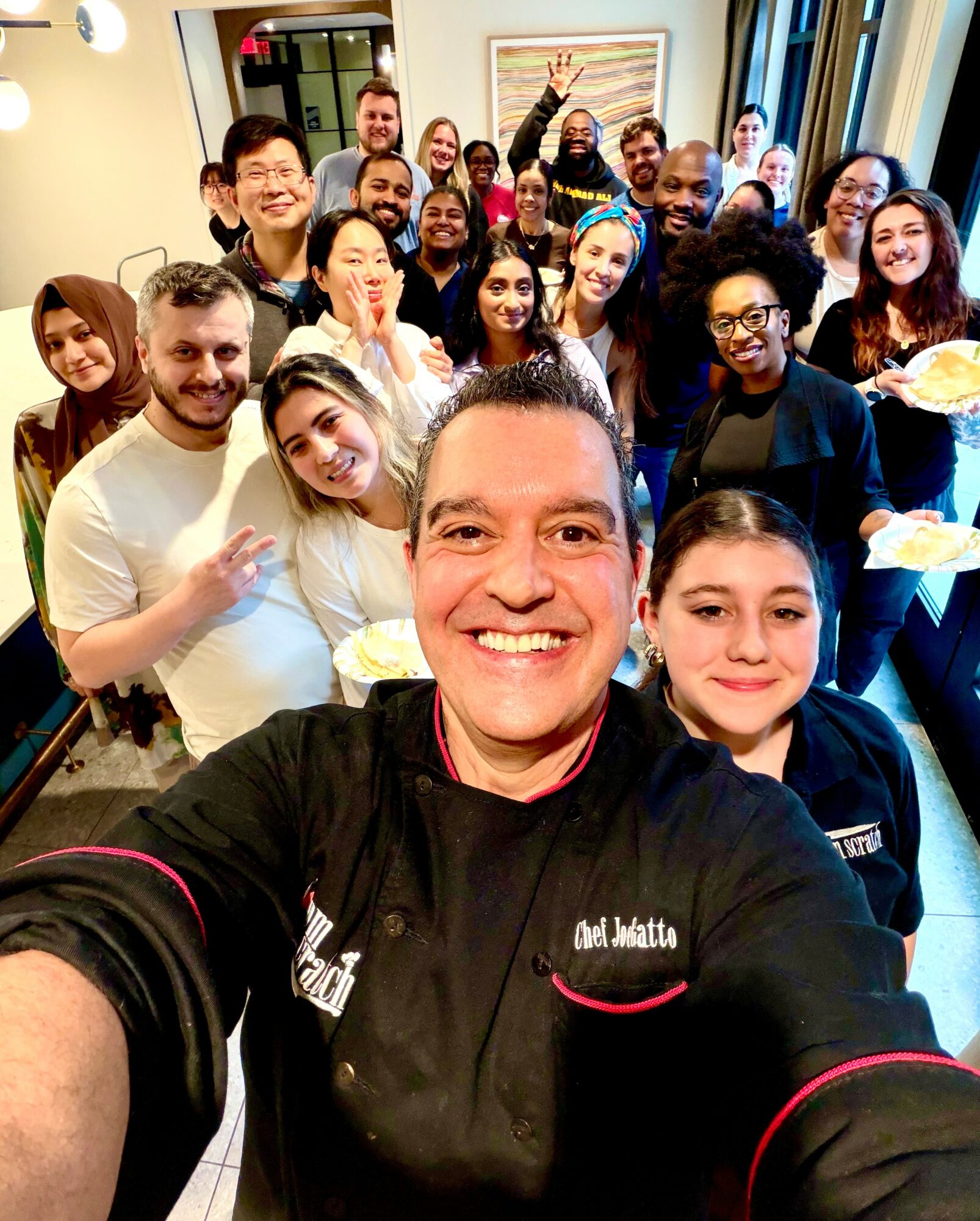 Group of diverse people smiling and holding plates in a room, with a person taking a selfie in the foreground.