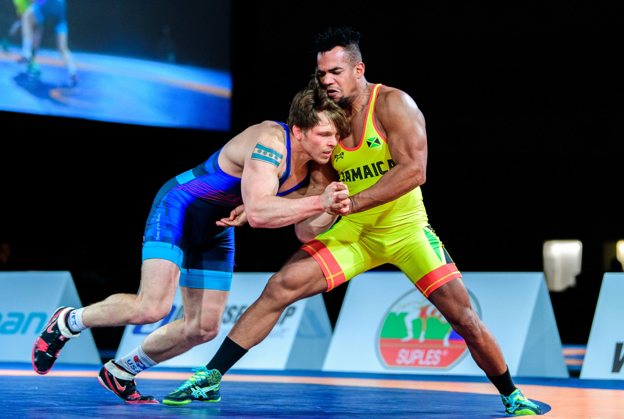 Two wrestlers compete on a blue mat, one in blue and the other in yellow and red, in a sports arena.