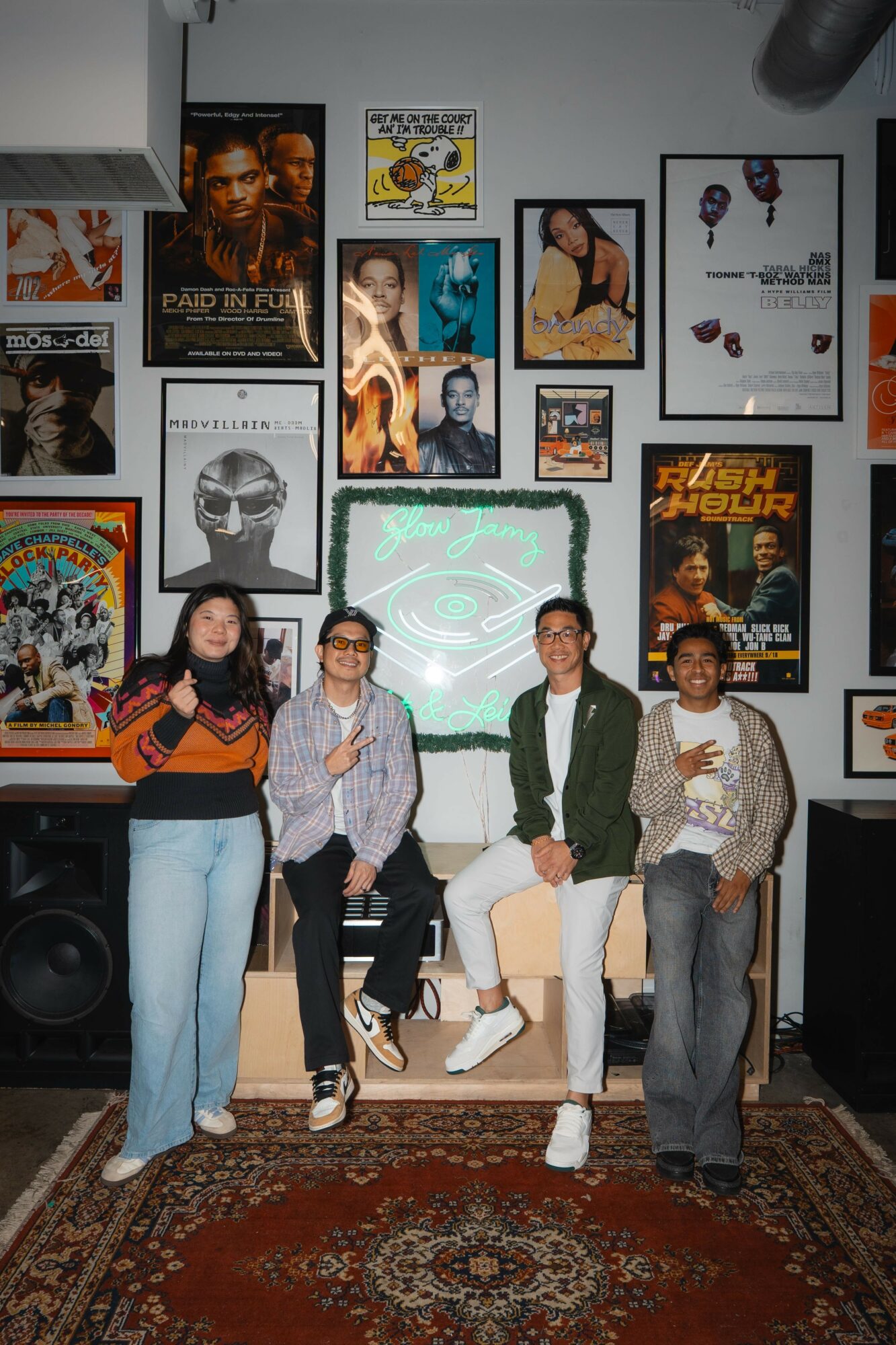 Four people standing in front of a wall with framed posters and artwork, some holding drinks, in a room with a patterned rug.