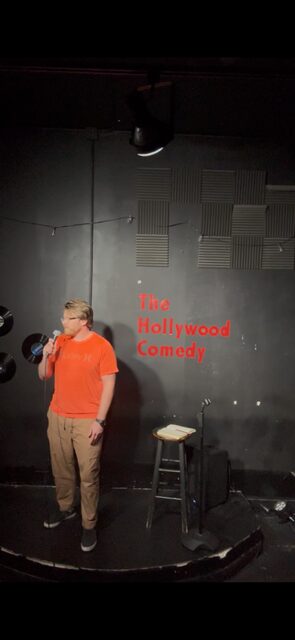 Person in orange shirt standing on stage with a stool and black box, gray wall background with red text.