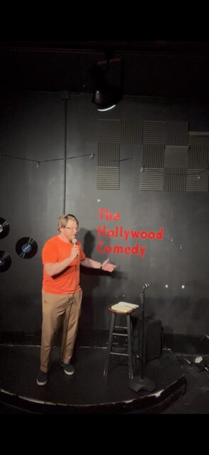 Stand-up comedian performs on stage with a microphone, stool, and a black wall background, with red text on the wall.