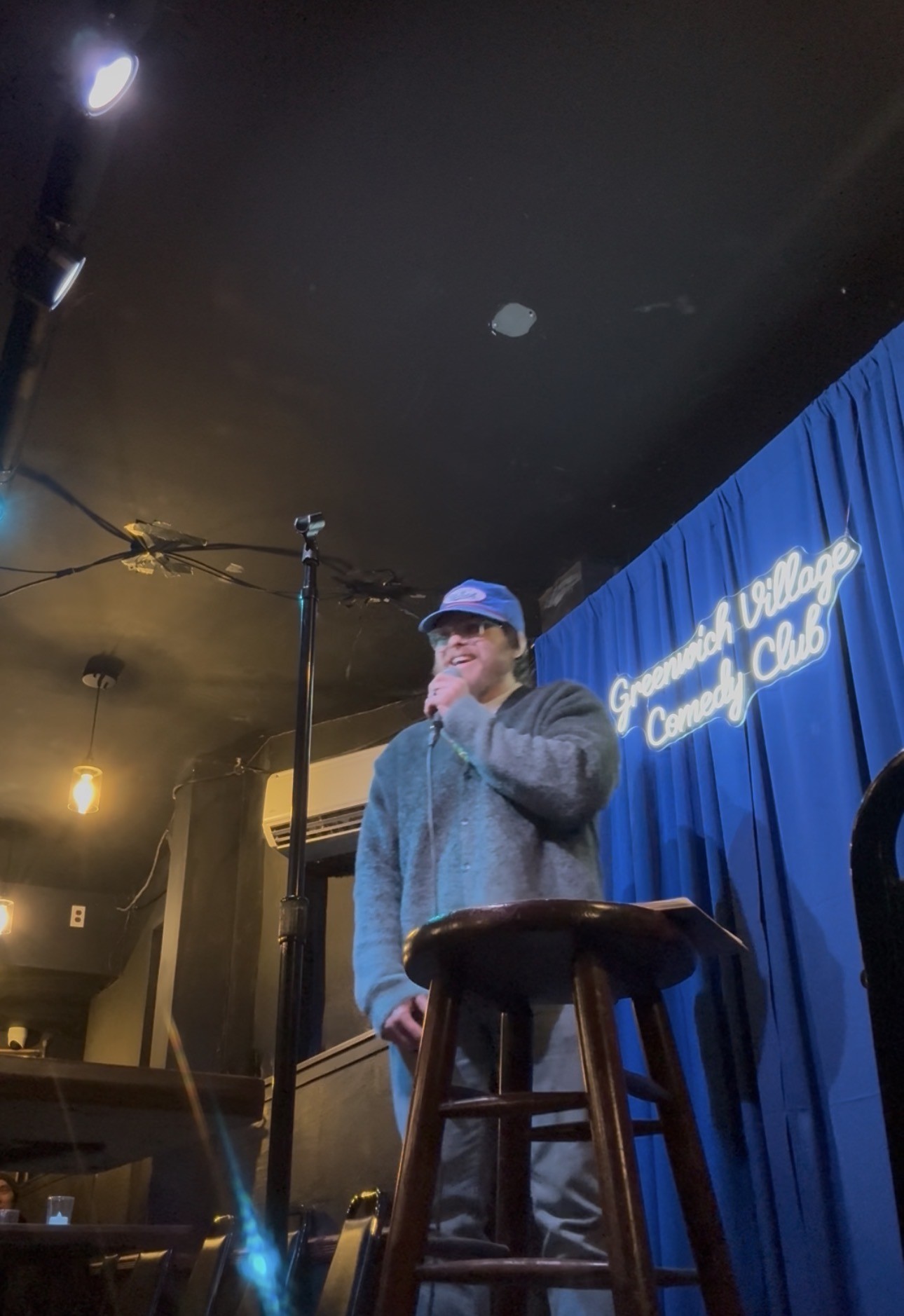 Person wearing a cap and sweater standing on stage with a blue curtain background and a stool nearby.