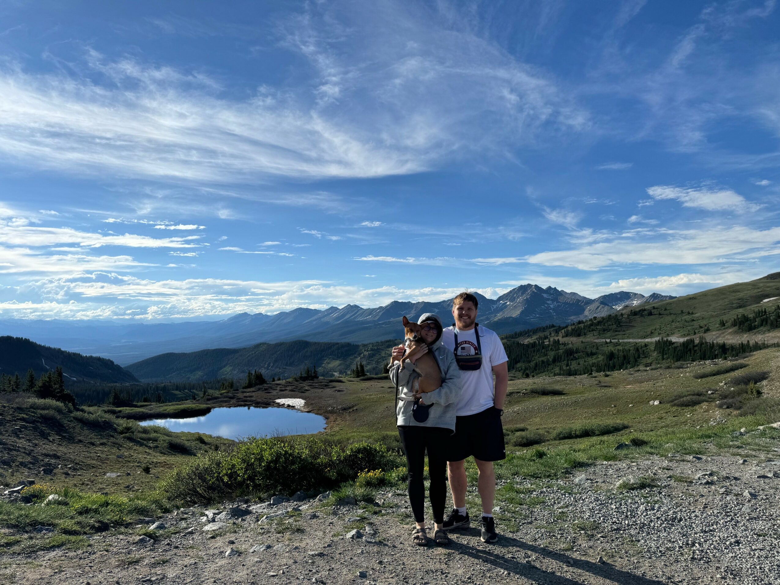 Two people standing on a grassy landscape with mountains, a lake, and a blue sky with clouds in the background.