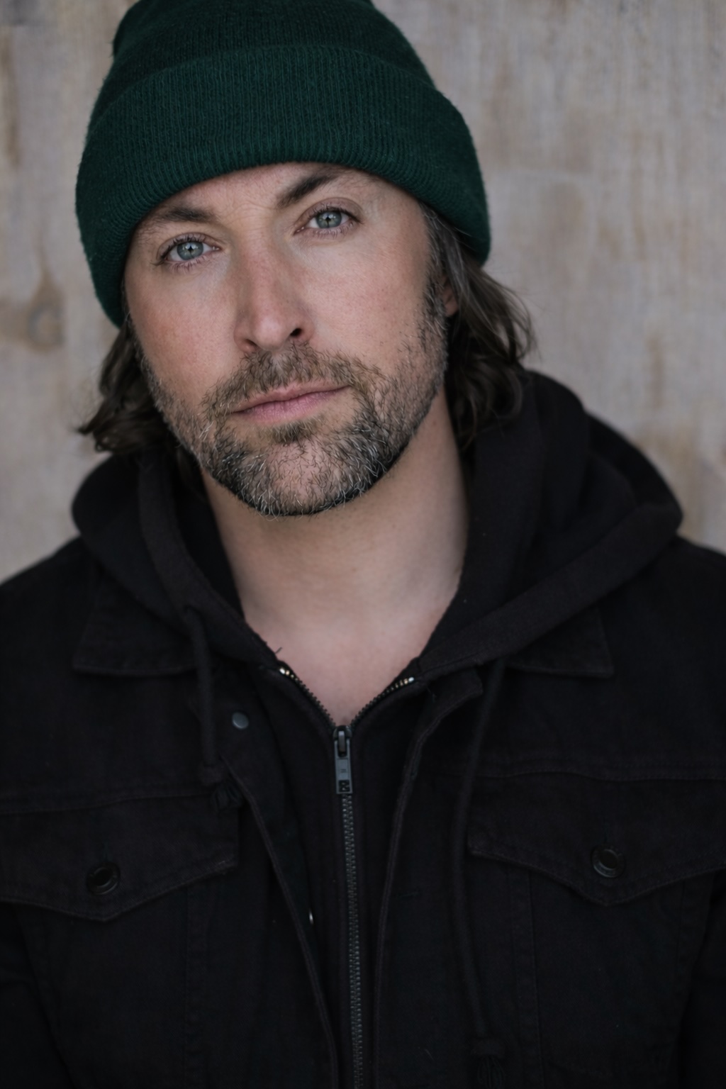 Man with dark hair, beard, wearing a green beanie and black jacket, looking at camera.