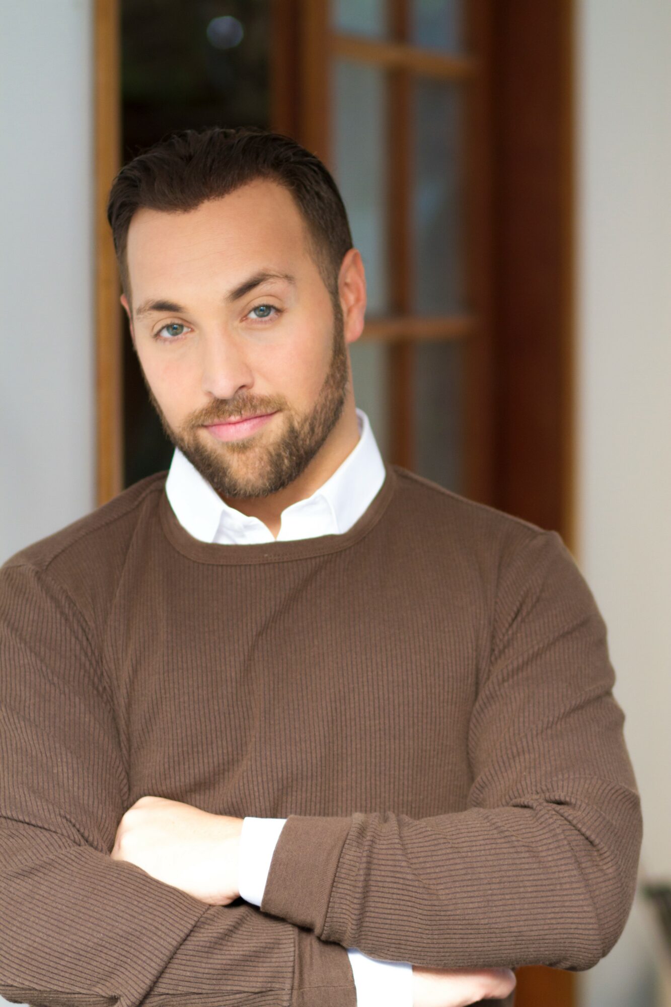 Man with dark hair and beard wearing a brown sweater and white shirt, arms crossed, indoors.