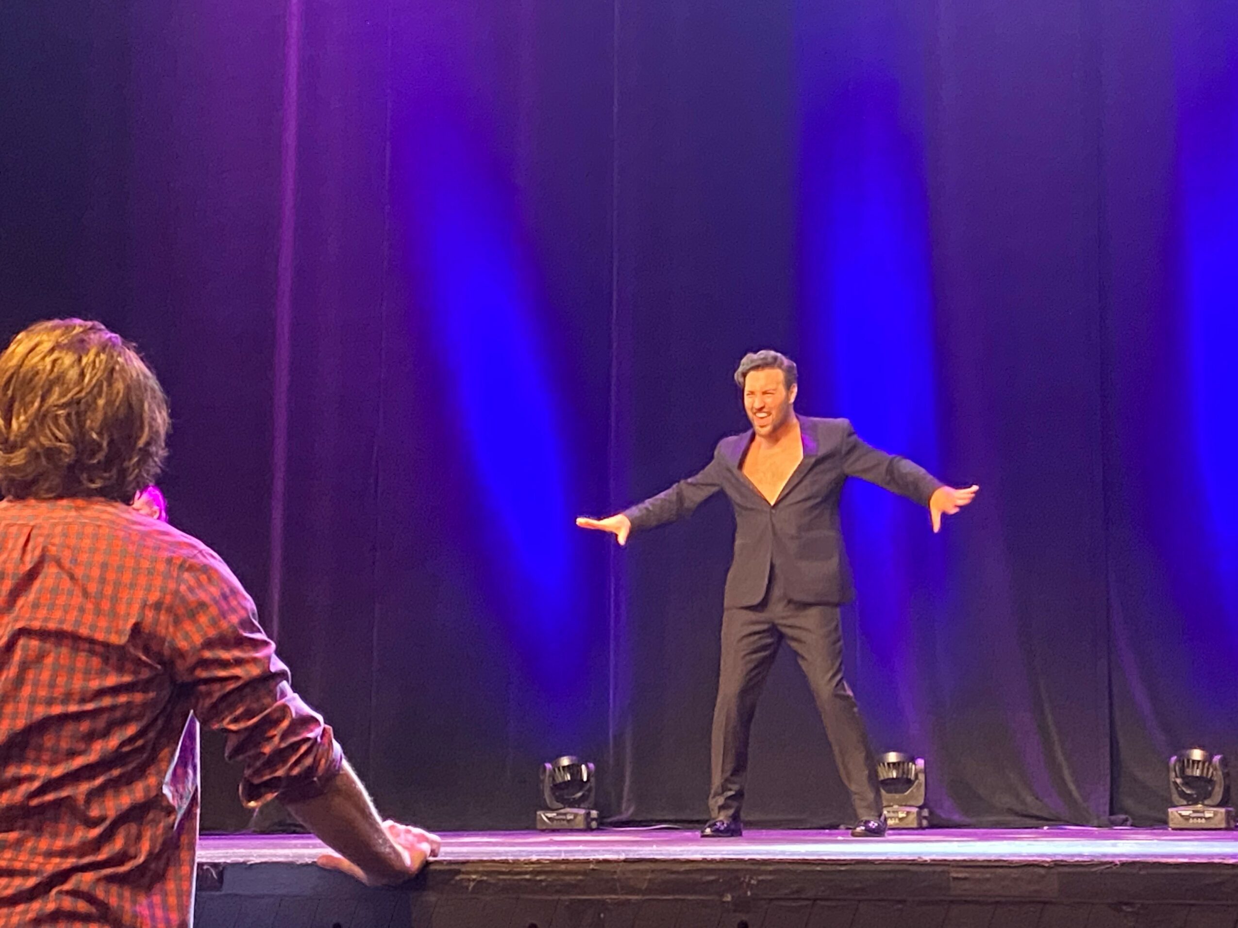 Person on stage with arms outstretched, facing another person in front, against a dark curtain backdrop with blue lighting.