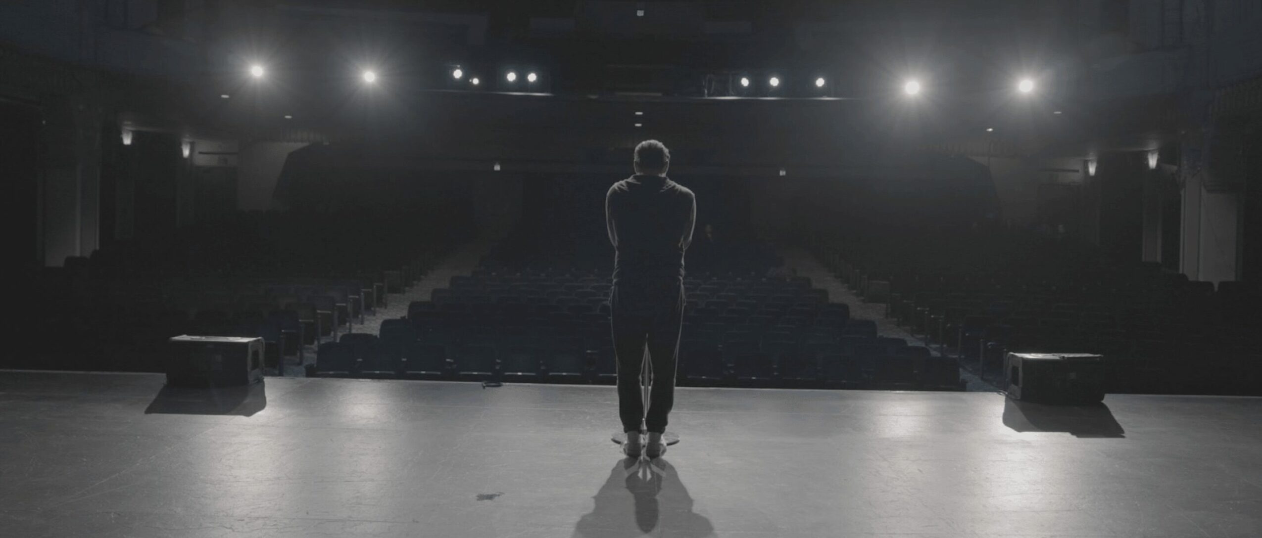 Person standing alone on stage with bright lights behind, empty auditorium in background.