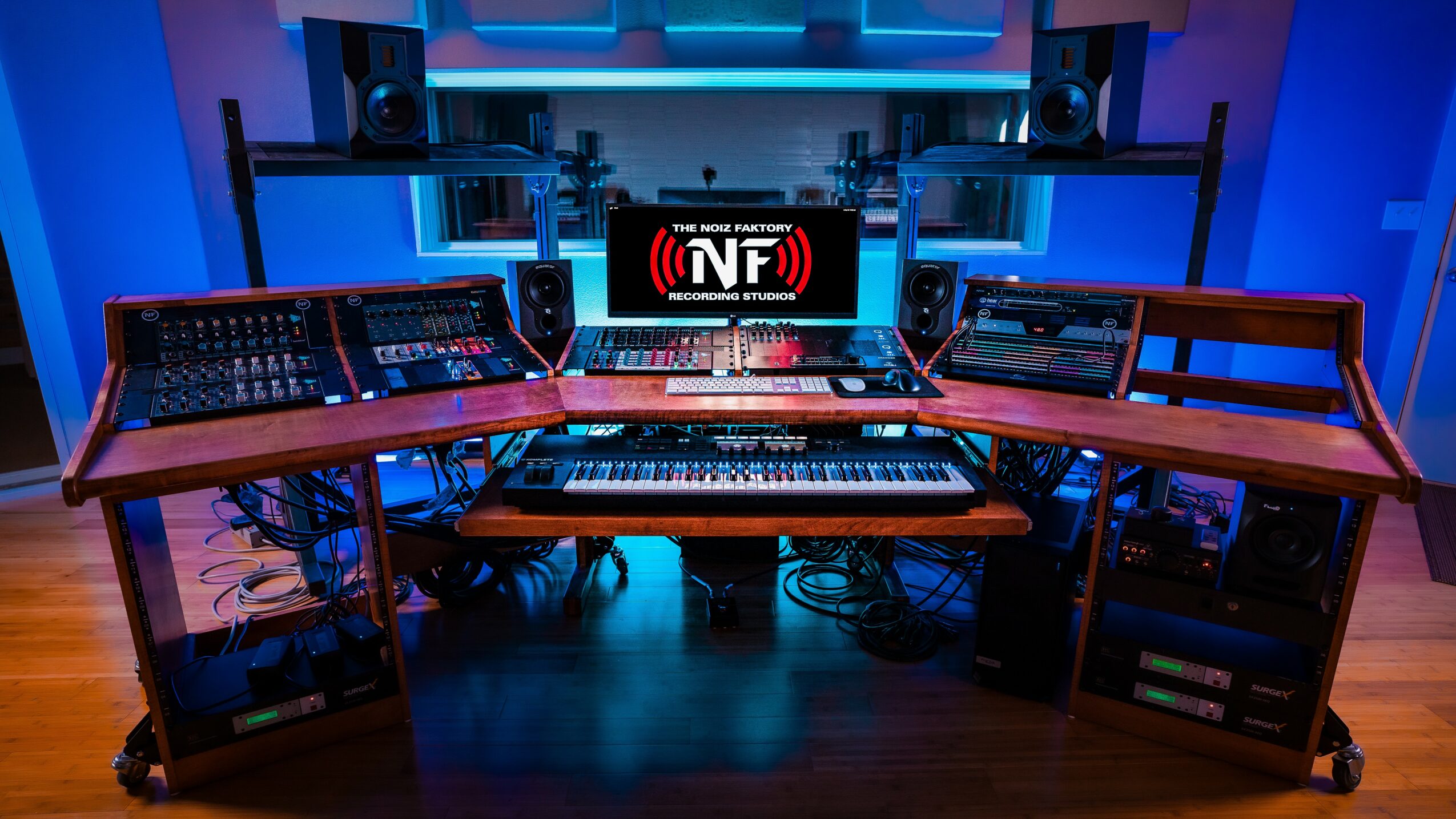 Music studio with mixing consoles, monitors, and electronic equipment on desks in a blue-lit room.