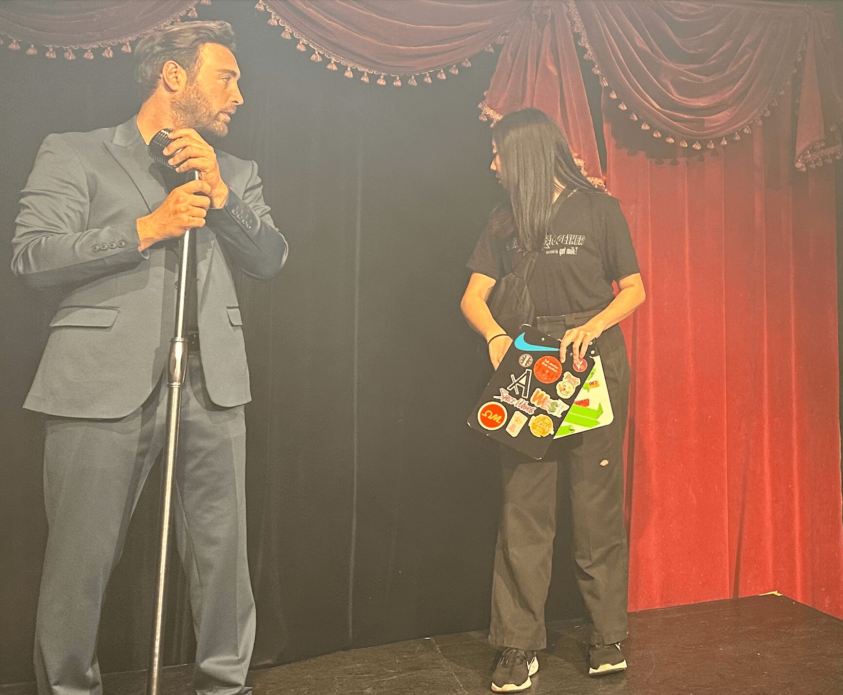 Man in suit holding microphone and woman in casual clothing holding a colorful box on stage with red curtains.