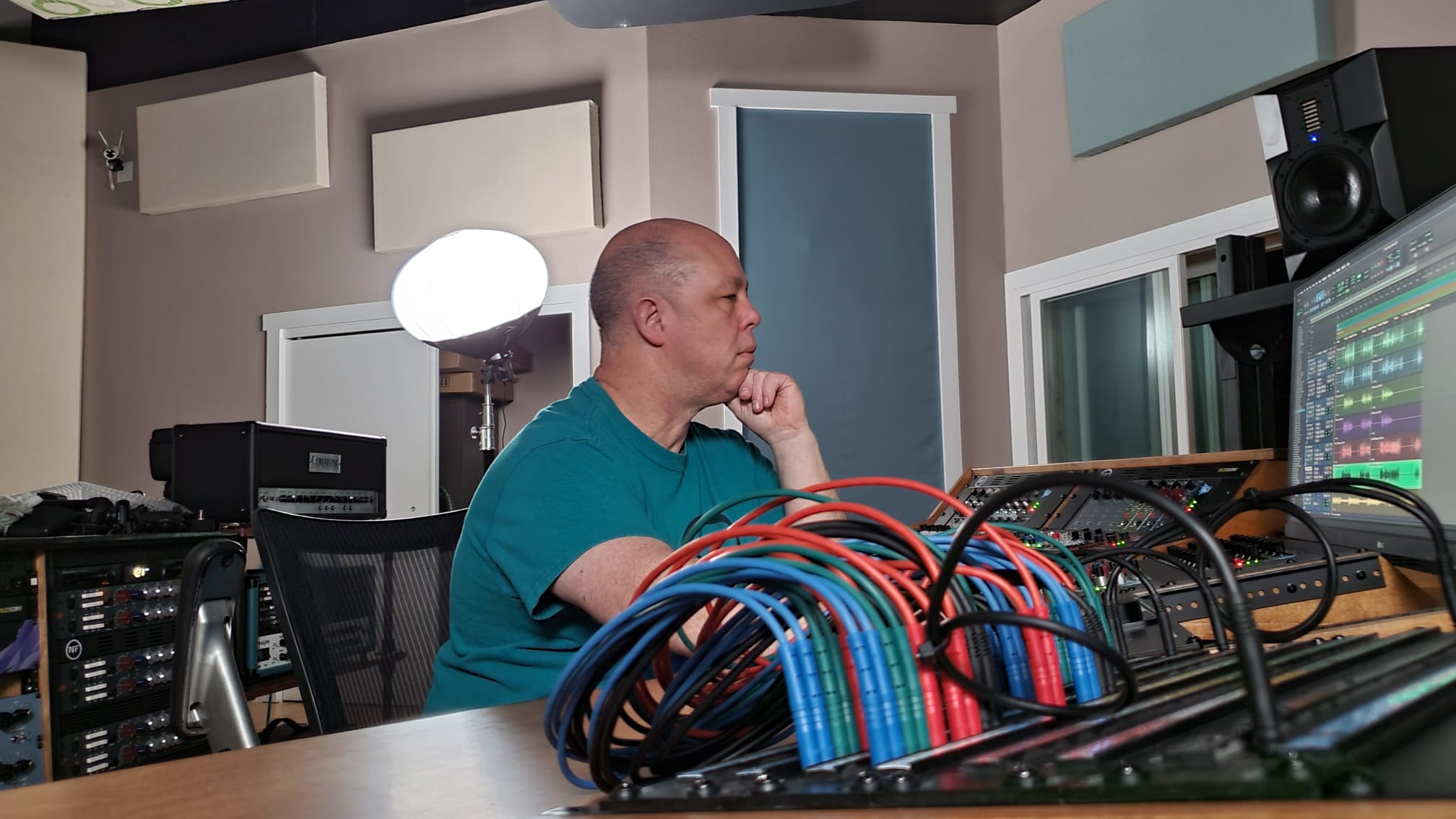 Person with bald head and teal shirt sitting at a desk in a recording studio with audio equipment and computer monitors.