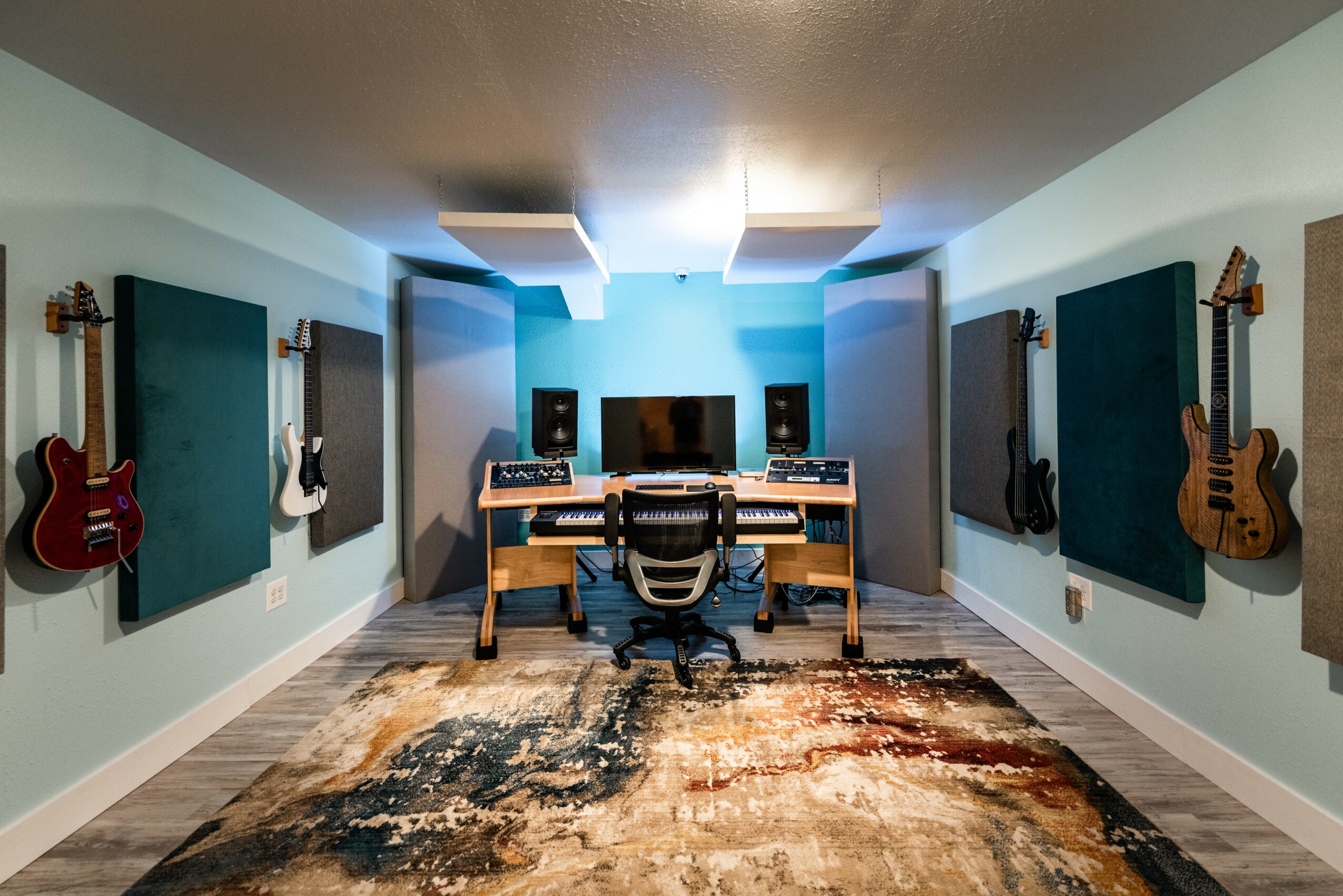 Music studio with guitars on walls, mixing console, monitors, and a central desk with a chair, on a colorful rug.
