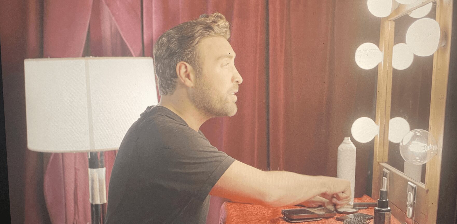 Man with short hair and beard sitting at a table with makeup products, in front of a mirror with lights.
