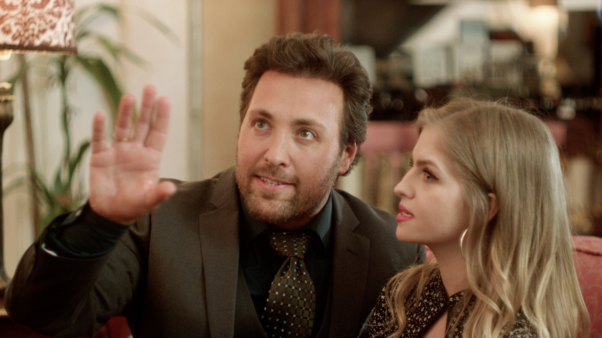 Man with dark hair and beard raising hand, woman with blonde hair looking at him, indoor setting.
