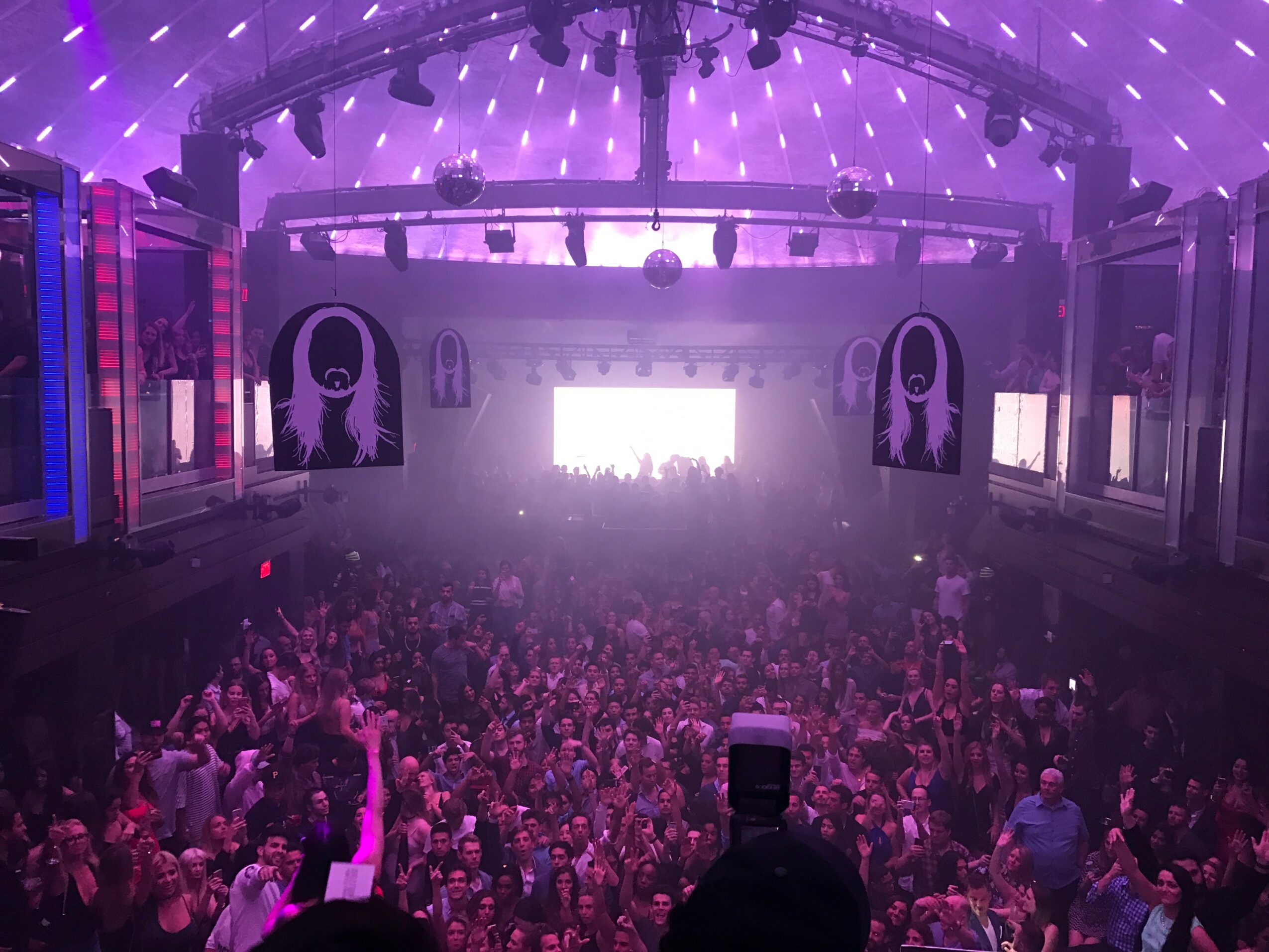 Crowd gathered in a large indoor concert venue with purple lighting and stage at the front.