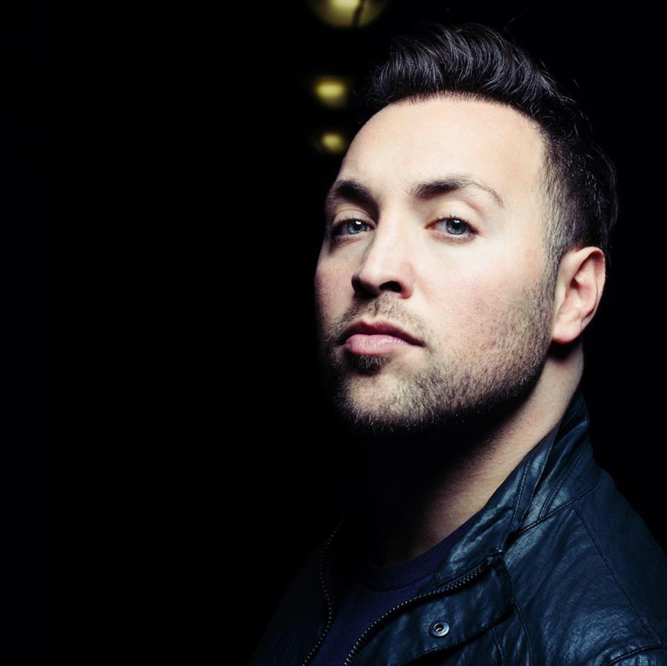 Portrait of a man with dark hair and a serious expression, wearing a dark leather jacket, against a dark background.