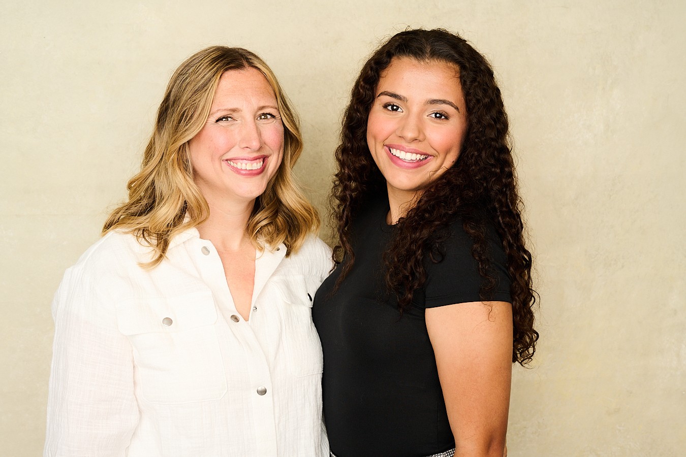 Two women smiling, standing close together against a plain background.