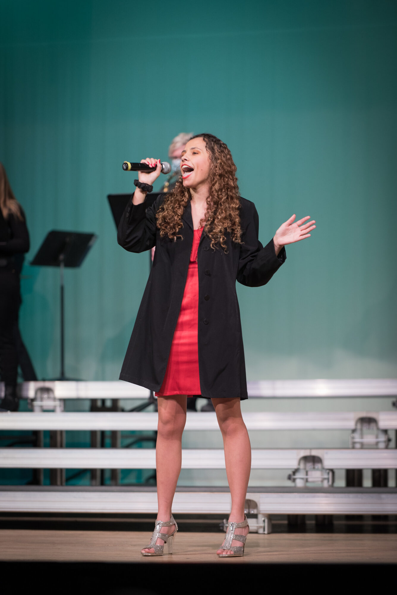 Young woman singing on stage with microphone, wearing red dress and black coat, with green backdrop.