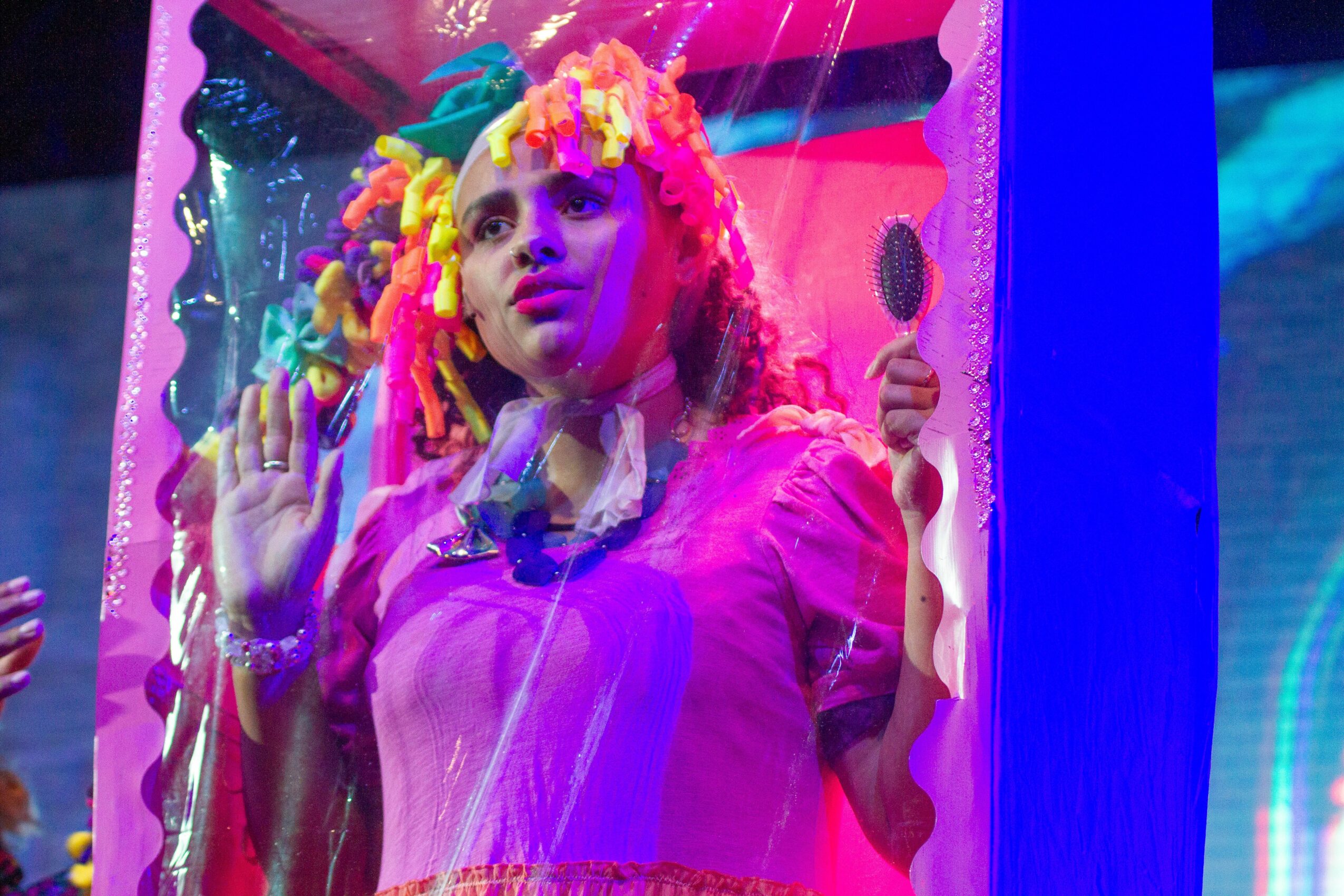 Woman with colorful dreadlocks looking through glass at a colorful display, holding a spoon, surrounded by vibrant colors.