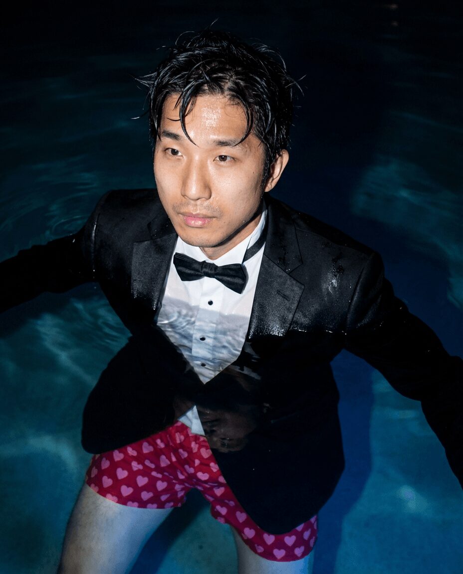 Man in tuxedo and bow tie in water, wearing pink polka dot shorts, looking to the side.