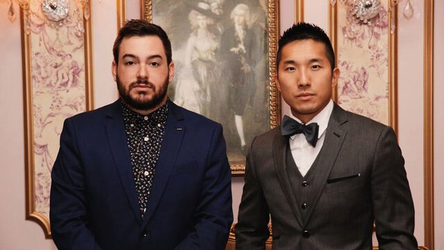 Two men in formal suits standing indoors with ornate wallpaper and framed painting behind them.