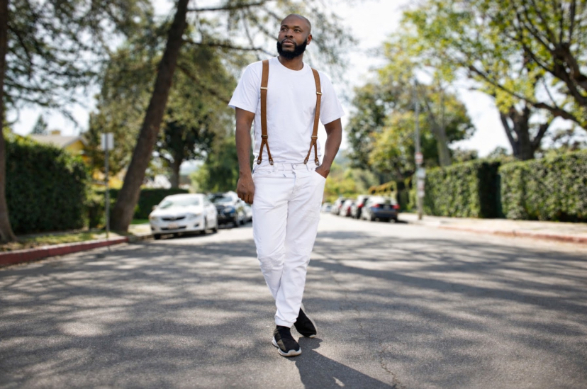 Man walking on street with trees and parked cars in background, wearing white outfit and carrying a backpack.