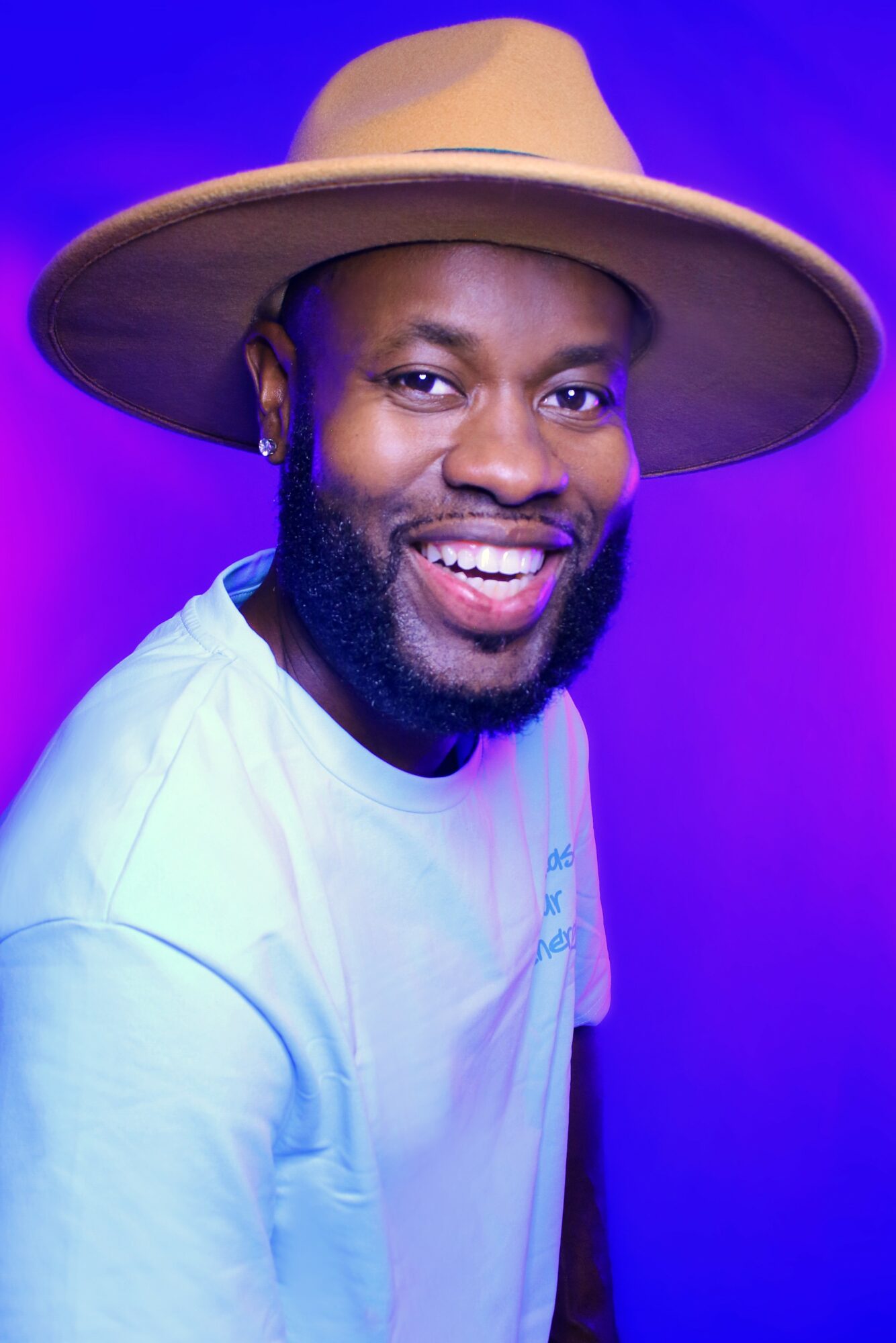 Smiling man wearing a wide-brimmed hat and white shirt against a purple background.