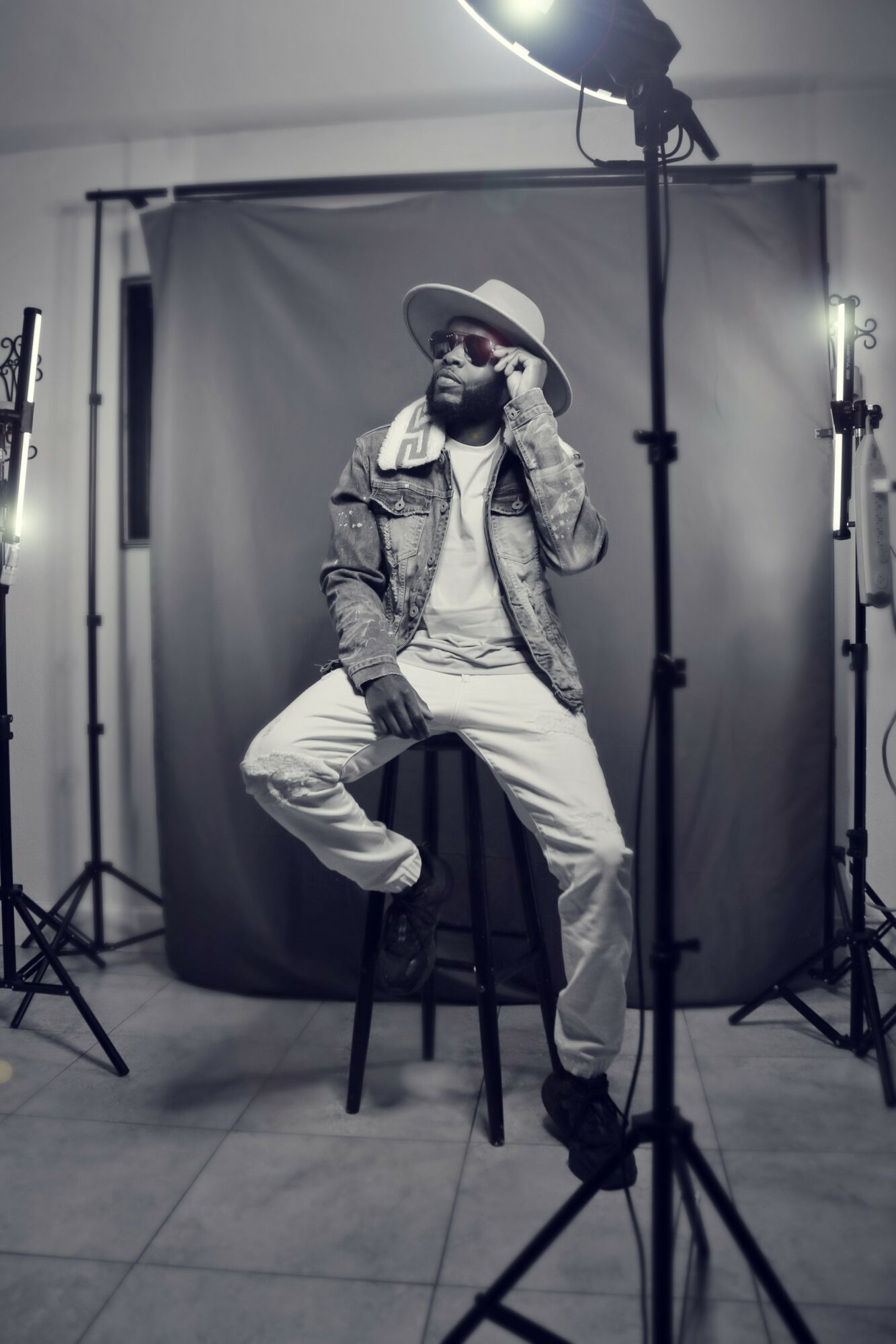 Person sitting on a stool in a photo studio, wearing a hat, sunglasses, and casual clothing, with studio lights and backdrop.